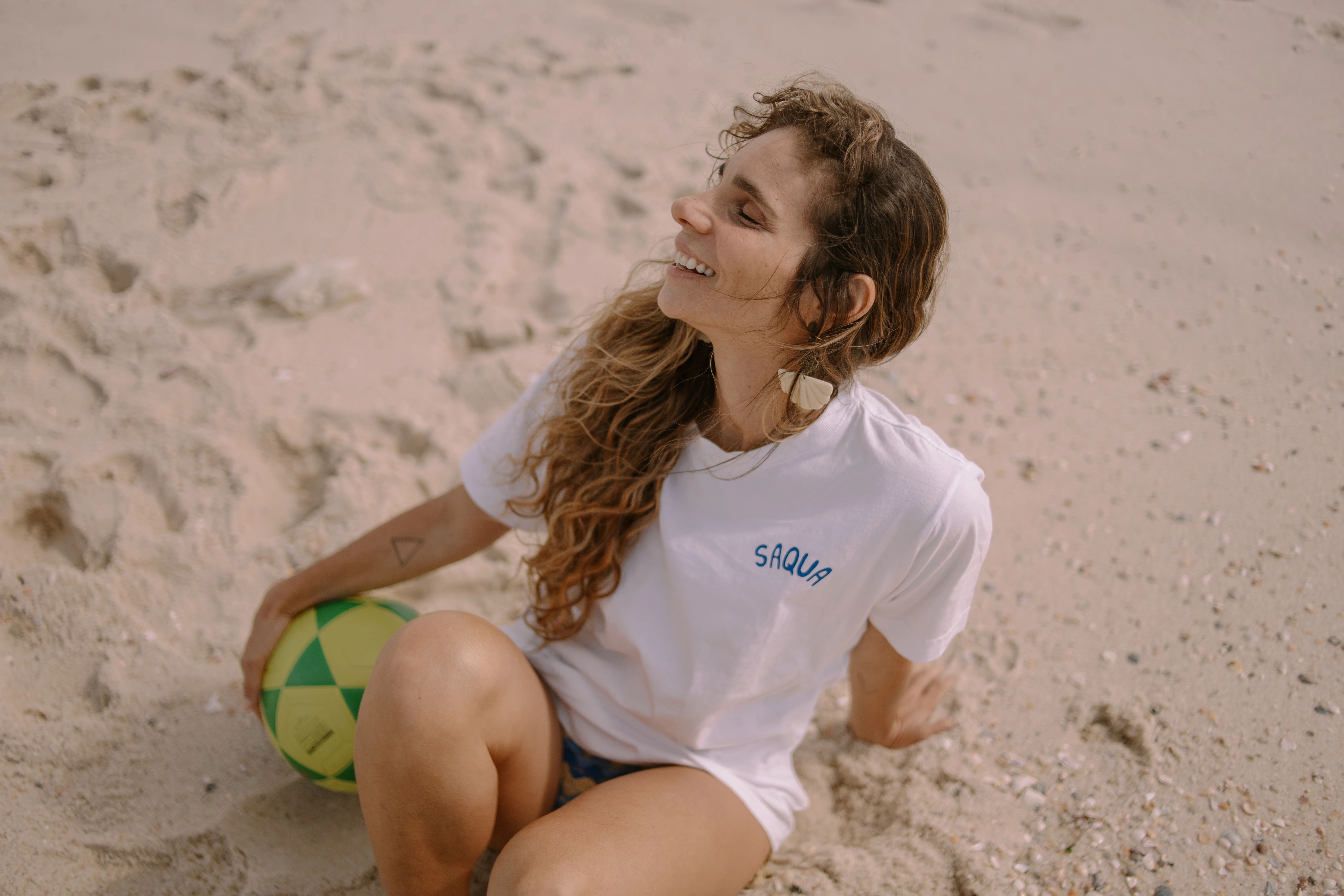 Modelo sorrindo sentada na areia da praia de Saquarema, com camiseta branca com o logo Saqua Vibes em azul e uma bola de futebol ao lado.
