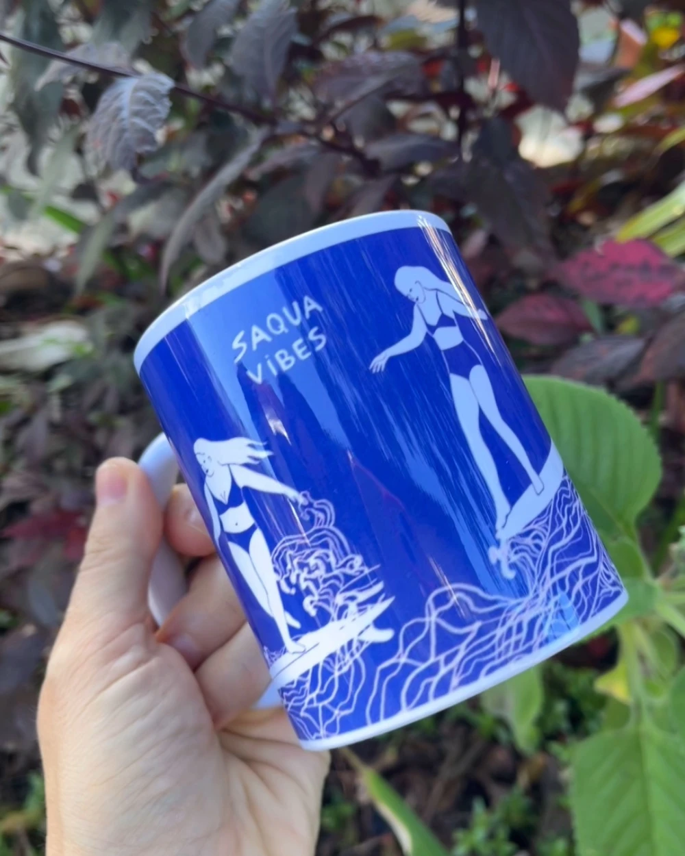Mão segurando a caneca azul com as surfistas da Saqua Vibes, entre folhagens.
