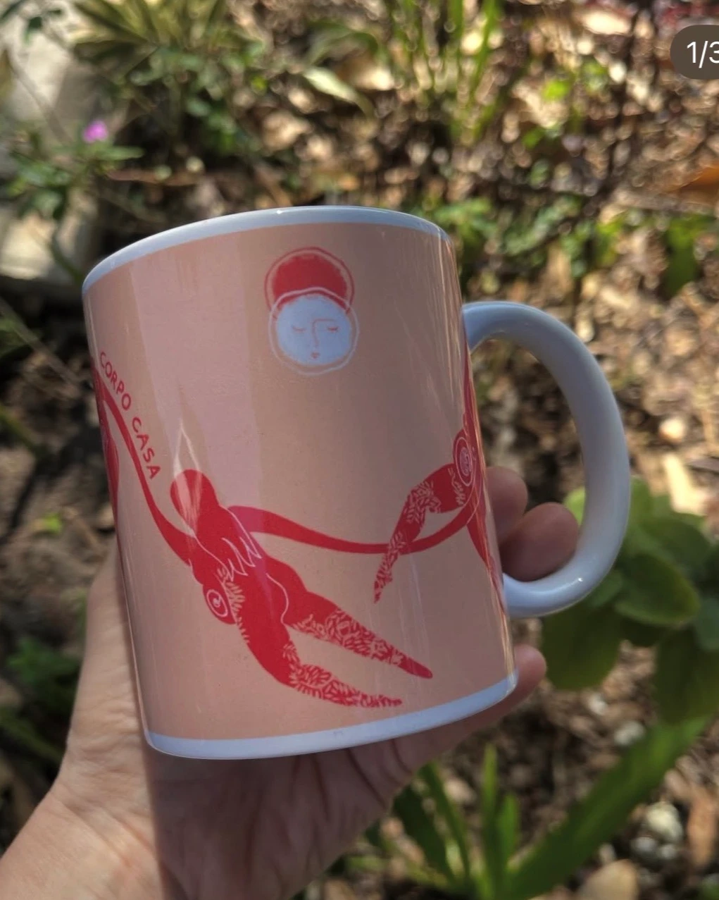 Mão segurando a caneca rosa Corpo Casa, com figuras vermelhas desenhadas a mão.