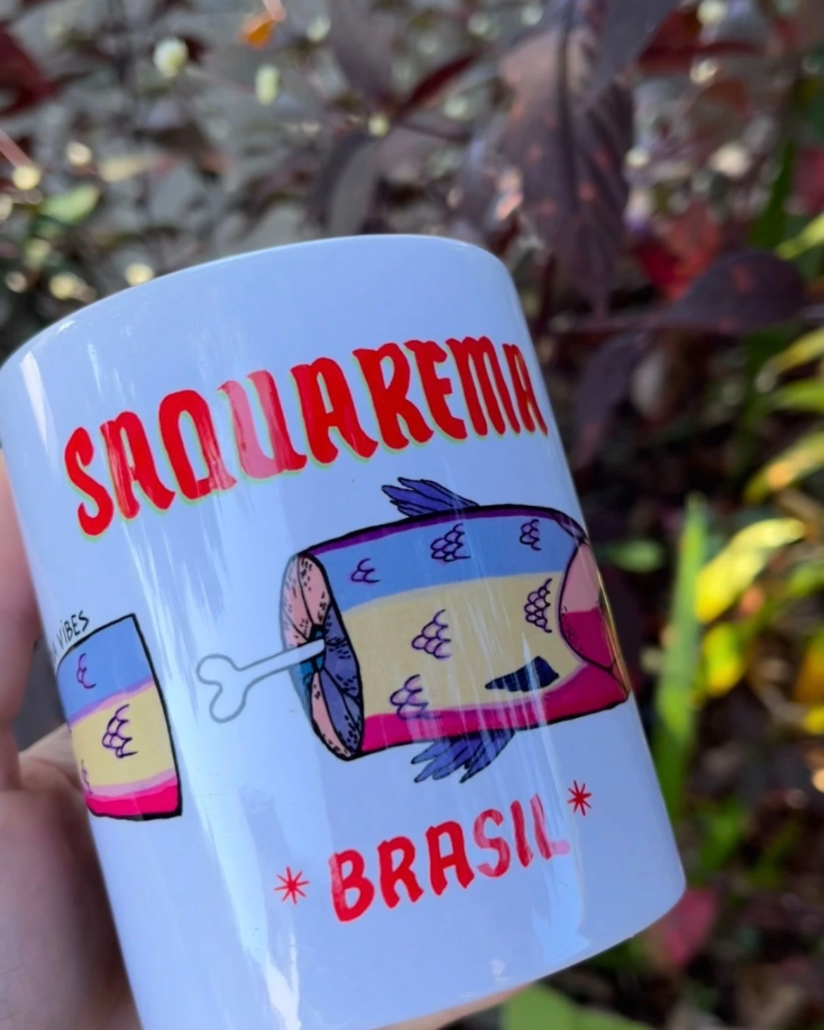 Mão segurando a caneca branca com a ilustração de um peixe e as palavras Saquarema Brasil.