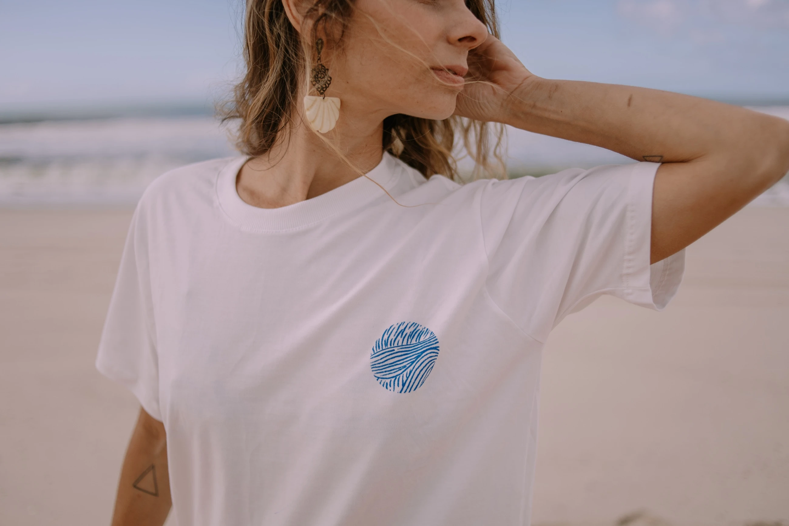 Mulher na praia com a mão no cabelo, vestindo camiseta branca com a estampa circular de ondas.