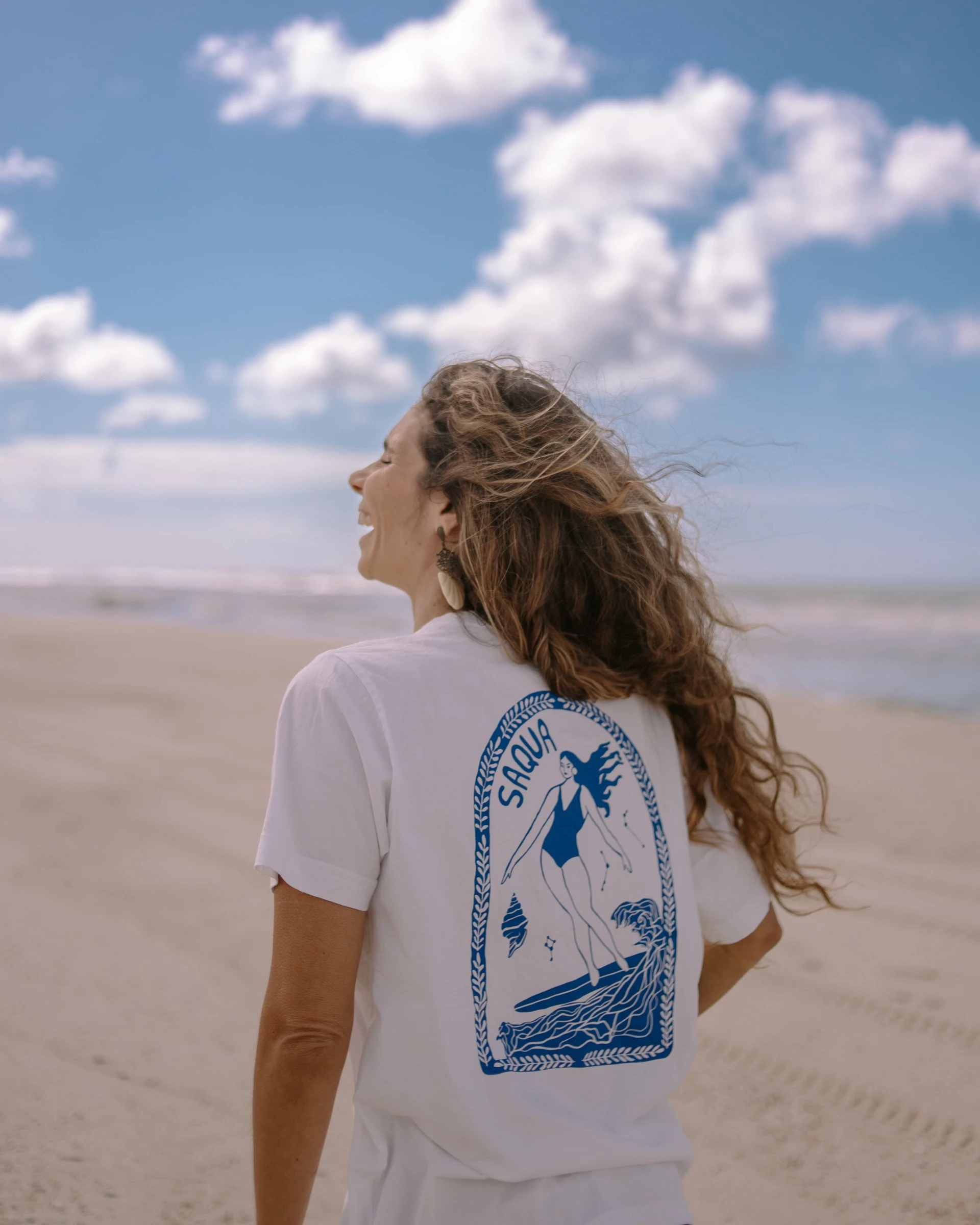 Mulher de costas na praia, rindo com o vento no cabelo, com a estampa do arco da surfista nas costas.