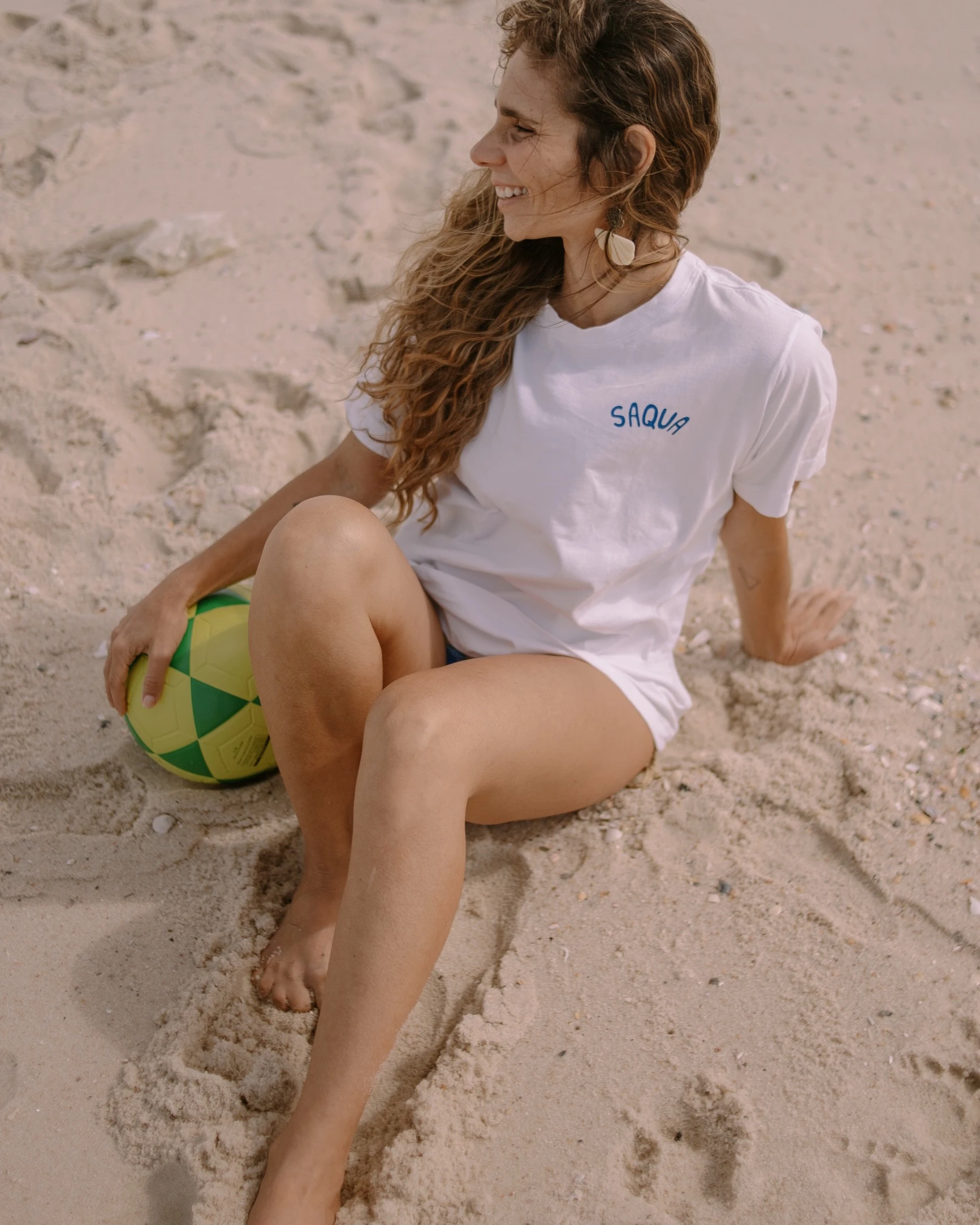 Mulher sentada na areia, sorrindo, com a camiseta da marca e uma bola nas mãos.