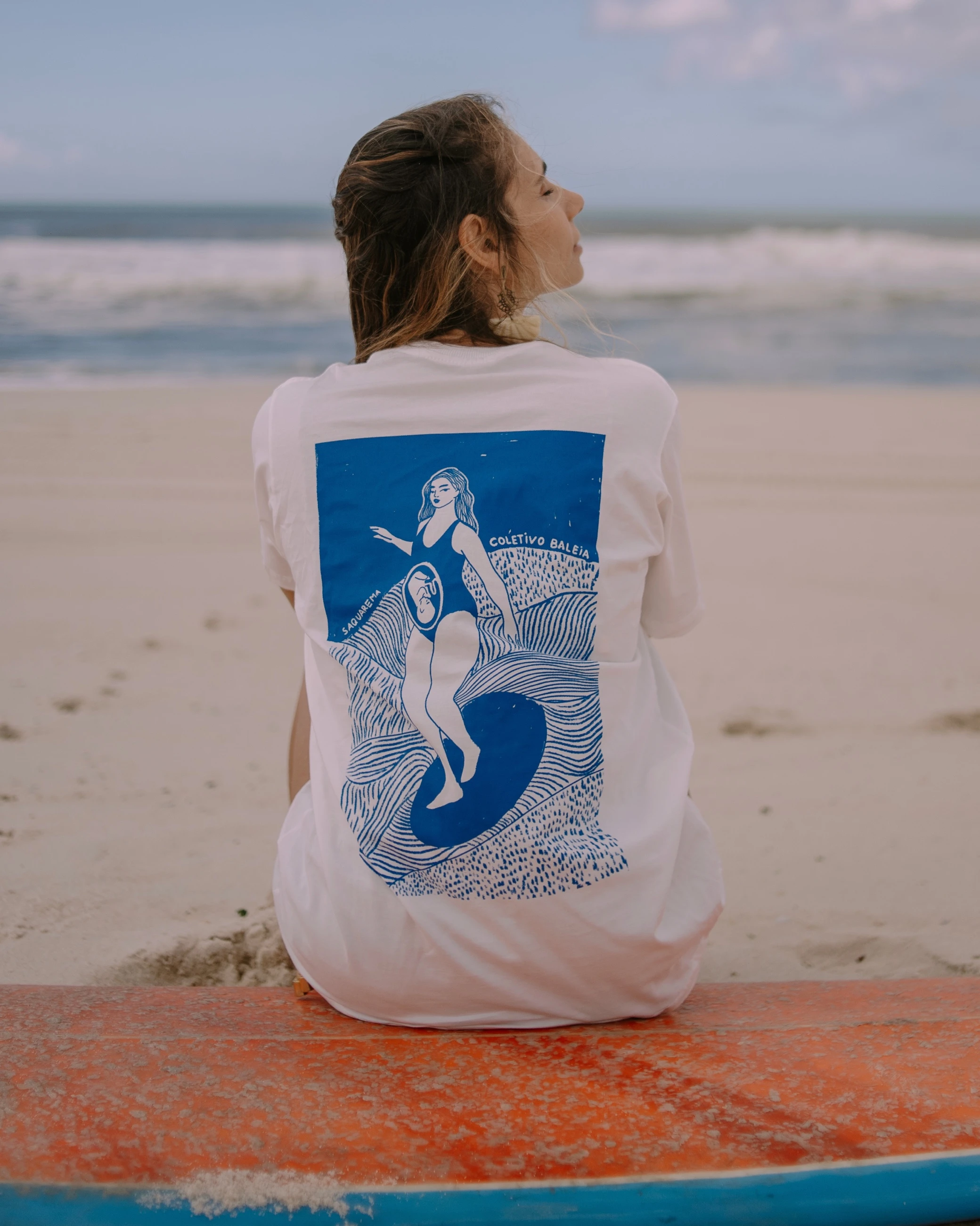Mulher sentada numa prancha, de costas, com a estampa grande de surfista nas costas.