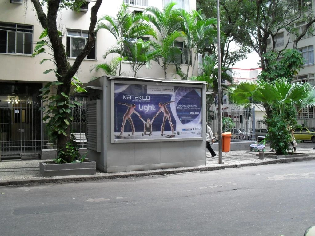 Painel do espetáculo Light, da Kataklò, em mobiliário urbano numa rua arborizada do Rio.